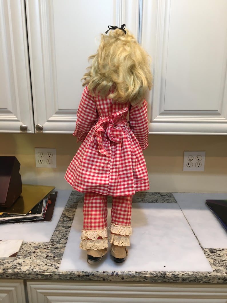 Peut inclure: Poup&eacute;e vintage v&ecirc;tue d'une robe &agrave; carreaux rouges et blancs et d'un pantalon assorti. La poup&eacute;e a des cheveux blonds et un n&oelig;ud noir. Le pantalon a une bordure en dentelle aux chevilles. La poup&eacute;e est sur une surface blanche.