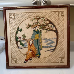 May include: A framed embroidery depicting two women in traditional Japanese clothing standing in a garden setting. The embroidery is done in a variety of colors, including green, blue, pink, and yellow. The frame is made of bamboo and has a dark brown finish.