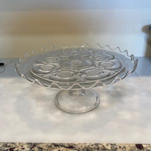 May include: A clear glass cake stand with a scalloped edge and a pedestal base. The stand has a decorative pattern of circles and dots.