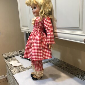 Peut inclure: Une poup&eacute;e vintage aux cheveux blonds boucl&eacute;s, v&ecirc;tue d'une robe &agrave; carreaux rouges et blancs, d'un pantalon assorti et de chaussures noires. La poup&eacute;e a un col en dentelle blanche et une ceinture rouge. La poup&eacute;e est debout sur une surface blanche.