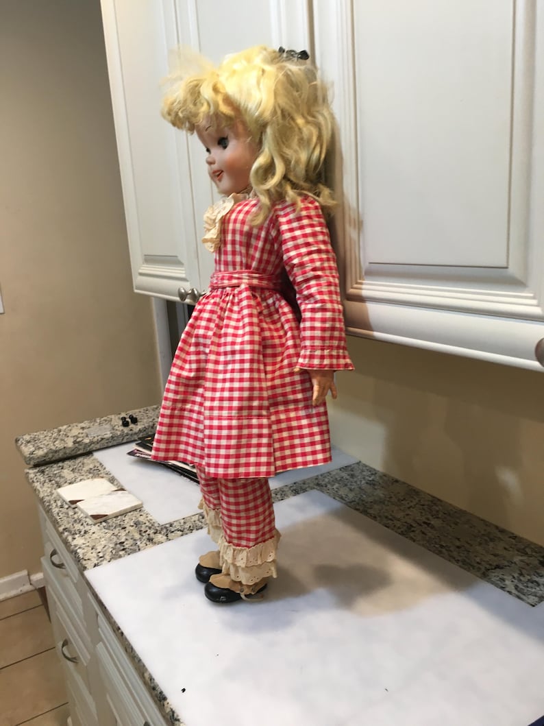 Peut inclure: Une poup&eacute;e vintage aux cheveux blonds boucl&eacute;s, v&ecirc;tue d'une robe &agrave; carreaux rouges et blancs, d'un pantalon assorti et de chaussures noires. La poup&eacute;e a un col en dentelle blanche et une ceinture rouge. La poup&eacute;e est debout sur une surface blanche.