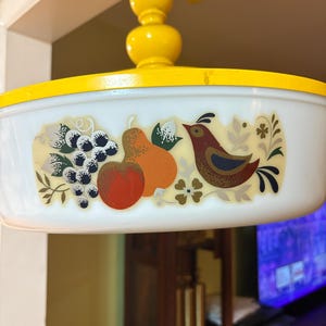 May include: Vintage white and yellow casserole dish with a lid. The dish features a colorful design of fruit and a bird. The lid and handle are bright yellow. A classic piece for serving and cooking.