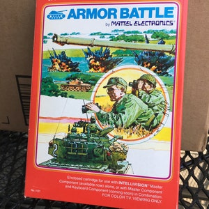 May include: A red and yellow box for the Intellivision video game "Armor Battle" by Mattel Electronics. The box features a cartoon illustration of a tank with soldiers and explosions. The text "FOR COLOR T.V. VIEWING ONLY" is printed on the bottom of the box.
