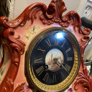 May include: A red and gold porcelain mantel clock with a black face and Roman numeral hour markers. The clock face has a small painted scene of a building and a river. The clock is decorated with floral designs.
