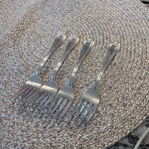 May include: A set of four silver-toned metal forks arranged in a row. Each fork features a slender handle with a subtle vertical grooved pattern and a rounded, decorative tip. The tines are polished and smooth, reflecting light across the surface.