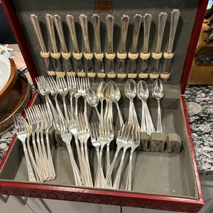 May include: A silver-toned flatware set housed in a dark red wooden storage case with a grey felt interior. The collection includes multiple knives, forks, and spoons arranged in designated slots, featuring a simple, polished metal design.