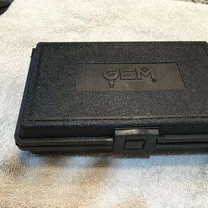 May include: Black rectangular case with the embossed word "GEM" on the lid. The case has a textured surface and a black latch closure. The case is closed and appears to be made of plastic.