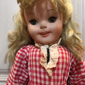 Peut inclure: Une poup&eacute;e vintage aux cheveux blonds ondul&eacute;s et aux yeux verts. La poup&eacute;e porte une robe &agrave; carreaux rouges et blancs avec un col cr&egrave;me bord&eacute; de dentelle. Le visage de la poup&eacute;e est peint avec des joues roses et une expression souriante.