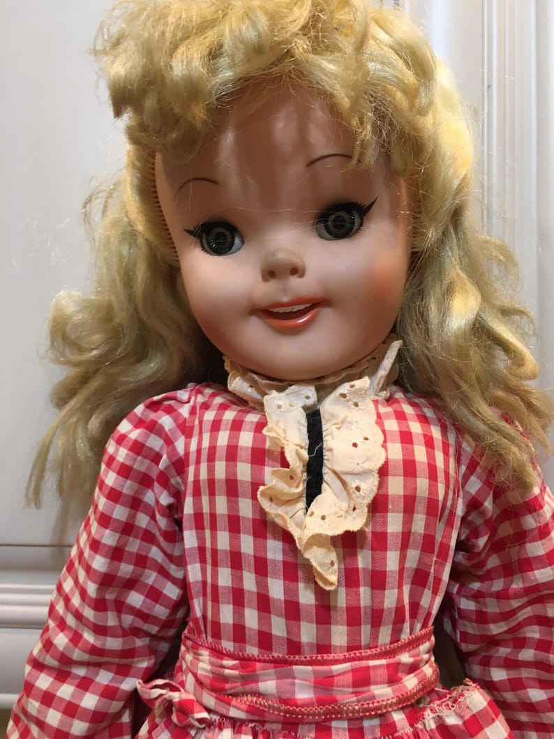 Peut inclure: Une poup&eacute;e vintage aux cheveux blonds ondul&eacute;s et aux yeux verts. La poup&eacute;e porte une robe &agrave; carreaux rouges et blancs avec un col cr&egrave;me bord&eacute; de dentelle. Le visage de la poup&eacute;e est peint avec des joues roses et une expression souriante.