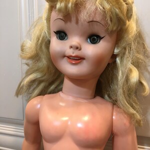 Peut inclure: Une poup&eacute;e vintage avec des cheveux blonds boucl&eacute;s. La poup&eacute;e a un visage peint avec de grands yeux verts, un eye-liner noir et une expression souriante. Le corps de la poup&eacute;e est fait d'un mat&eacute;riau de couleur p&ecirc;che clair.