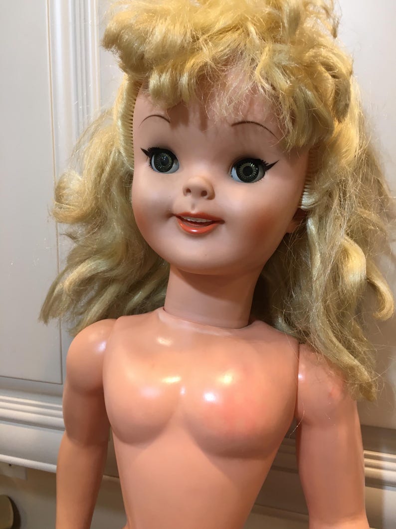 Peut inclure: Une poup&eacute;e vintage avec des cheveux blonds boucl&eacute;s. La poup&eacute;e a un visage peint avec de grands yeux verts, un eye-liner noir et une expression souriante. Le corps de la poup&eacute;e est fait d'un mat&eacute;riau de couleur p&ecirc;che clair.