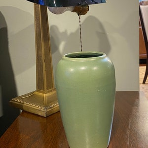 May include: A green ceramic vase with a wide mouth and a rounded body sits on a wooden table next to a vintage brass desk lamp with a green glass shade.