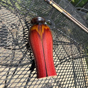 May include: A cylindrical wooden vessel with a smooth, glossy finish featuring a deep red and dark brown gradient. The object has a rounded top section and a slightly flared base, showcasing natural wood grain patterns beneath the polished surface.