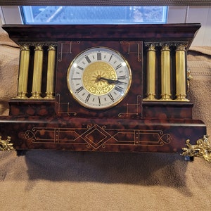 May include: A vintage-style mantel clock with a dark brown wood case and gold accents. The clock face has Roman numerals and a gold-colored center. The clock is sitting on a brown surface.