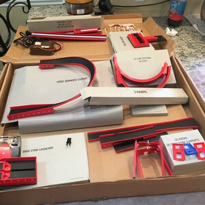 May include: A box of red and black plastic track pieces for a toy race car set. The pieces include a drag strip launcher, high banked curve, loop sections with clips, ramps, and racks and supports.