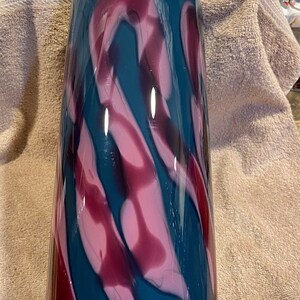 May include: A tall, cylindrical glass bottle with a teal base color. Swirls of pink and burgundy flow across the surface. The bottle has a shiny, reflective finish. The top of the bottle is a gold color.