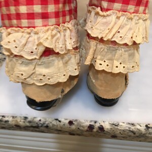Peut inclure: Gros plan sur les jambes d'une poup&eacute;e. Les jambes sont recouvertes d'un tissu &agrave; carreaux rouges et blancs, avec une garniture en dentelle cr&egrave;me autour des chevilles. Les chaussures de la poup&eacute;e sont de couleur beige avec des semelles noires.