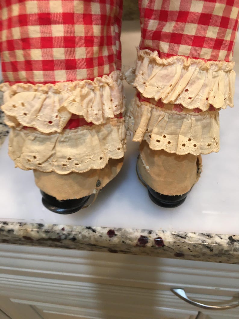 Peut inclure: Gros plan sur les jambes d'une poup&eacute;e. Les jambes sont recouvertes d'un tissu &agrave; carreaux rouges et blancs, avec une garniture en dentelle cr&egrave;me autour des chevilles. Les chaussures de la poup&eacute;e sont de couleur beige avec des semelles noires.