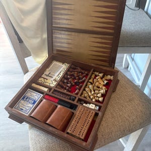 May include: A wooden game set in a hinged box featuring a backgammon board on the interior lid. The base contains compartments holding dark and light chess pieces, red and black checkers, two brown dice cups, dice, a cribbage board, and a deck of Lions playing cards.