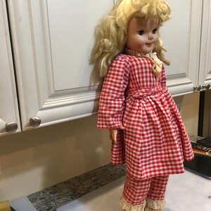 Peut inclure: Une poup&eacute;e vintage aux cheveux blonds boucl&eacute;s, v&ecirc;tue d'une robe &agrave; carreaux rouges et blancs et d'un pantalon assorti. La poup&eacute;e a un col et des poignets en dentelle blanche et porte des chaussures noires avec des bottes beiges. La poup&eacute;e est devant un placard blanc.