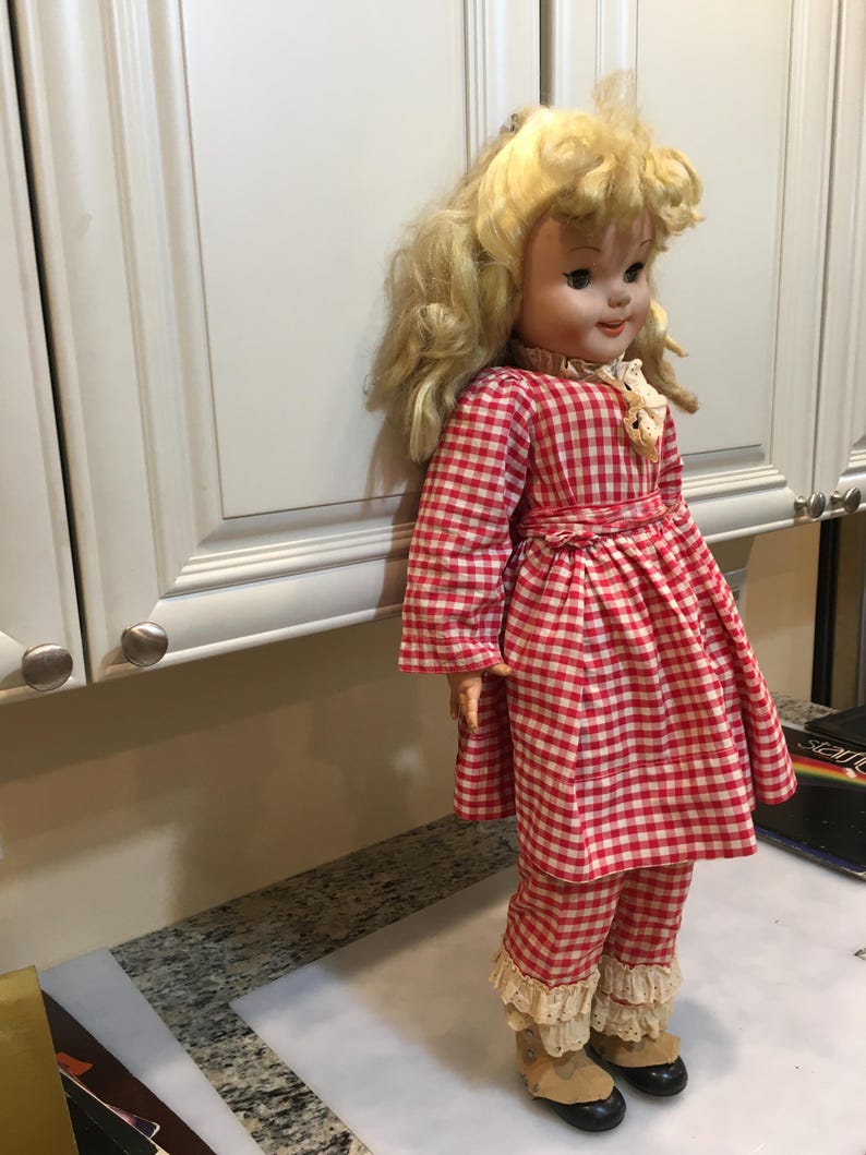 Peut inclure: Une poup&eacute;e vintage aux cheveux blonds boucl&eacute;s, v&ecirc;tue d'une robe &agrave; carreaux rouges et blancs et d'un pantalon assorti. La poup&eacute;e a un col et des poignets en dentelle blanche et porte des chaussures noires avec des bottes beiges. La poup&eacute;e est devant un placard blanc.