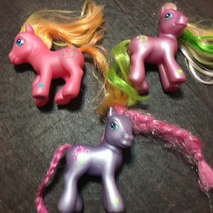 May include: Three small plastic pony toys with colorful hair. The ponies are pink, purple, and light pink. They have painted on eyes and a flower design.