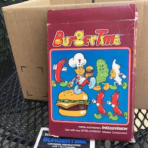 May include: A red and blue box for the Intellivision video game "BurgerTime". The box features a cartoon chef holding a burger, a pickle, and an egg. The text "BurgerTime" is in yellow letters on a red background. The text "MATTEL ELECTRONICS INTELLIVISION" is in white letters on a blue background. The text "Use with any INTELLIVISION Master Component" is in white letters on a blue background.