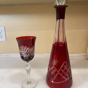Puede incluir: Una decantador de cristal rojo con un tapón de vidrio transparente y una copa de vino de cristal rojo a juego. La decantador y la copa tienen intrincados diseños cortados.