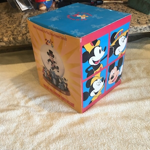 May include: A blue and yellow box with a Mickey Mouse snow globe inside. The box has a picture of Mickey Mouse on the front and the words "Mickey's Millennium Waterglobe" on the bottom. The box is made exclusively for May Department Stores.