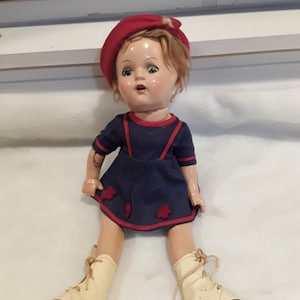 May include: A vintage doll wearing a red beret, a blue and white sailor dress, and white roller skates. The doll has blonde hair and blue eyes.