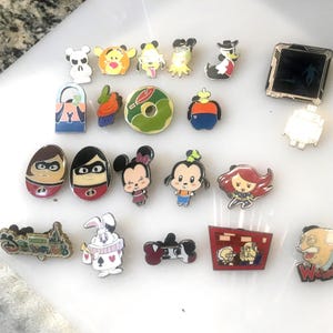 May include: A collection of colorful enamel pins featuring various cartoon characters and designs. The pins include characters from Disney, The Incredibles, and other animated series. The pins are in various shapes and sizes, with a glossy finish.