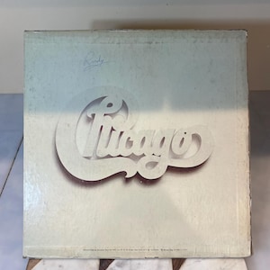 May include: A white vinyl record album cover with the word "Chicago" in a stylized font. The album cover has a handwritten signature that says "Rudy".