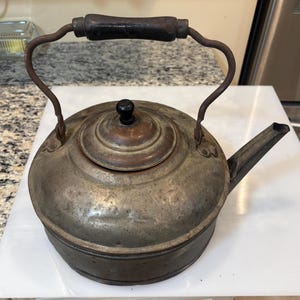 May include: Antique metal kettle with a dark wooden handle and a black knob on the lid. The kettle has a spout and a curved handle. The metal has a tarnished, aged appearance, suggesting it is vintage.