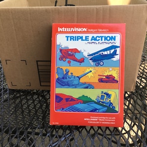 May include: A red, white, and blue game cartridge for the Intellivision Intelligent Television. The cartridge is labeled "Triple Action" by Mattel Electronics. The image shows three scenes from the game: a plane dogfight, a tank battle, and a car chase.