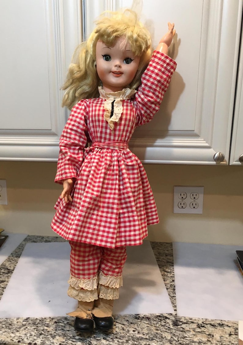 Peut inclure: Poup&eacute;e vintage aux cheveux blonds et aux yeux bleus, v&ecirc;tue d'une robe &agrave; carreaux rouges et blancs, d'un pantalon assorti et de chaussures noires. La poup&eacute;e a un col en dentelle blanche et est pos&eacute;e avec un bras lev&eacute;.