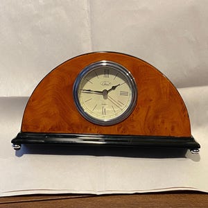 May include: A wooden clock with a black base and a silver face. The clock has a wood grain pattern and the face has Roman numerals.