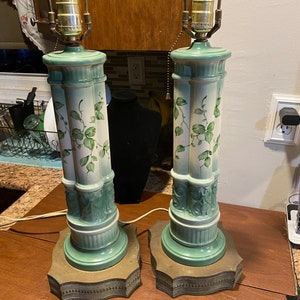 May include: A pair of vintage ceramic table lamps with green and white floral designs. The lamps have a columnar shape with a green base and a brass-colored bottom.
