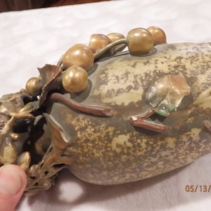May include: A ceramic vase with a brown and green glaze. The vase is decorated with a vine of gold-colored berries and leaves. The vase has a unique, organic shape.