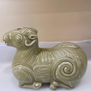May include: A green ceramic figurine of a ram with intricate details, including a spiral design on the back. The ram is sitting on a white surface.