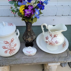 May include: A set of vintage white porcelain bathroom accessories with a red floral design. The set includes a pitcher, a bowl, a lidded jar, and a small soap dish.