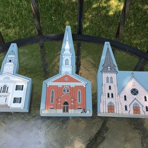 May include: Three wooden church building models. The first is white with green trim and a clock tower. The second is red brick with a white steeple. The third is light pink with a gray steeple and a round stained glass window.