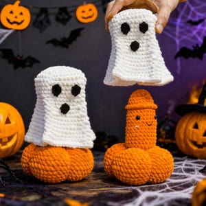 May include: Crocheted Halloween decorations featuring a ghost and a pumpkin-shaped figure. The ghost is white with black eyes, and the pumpkin figure is orange. The background includes pumpkins, spiders, and a witch's hat.