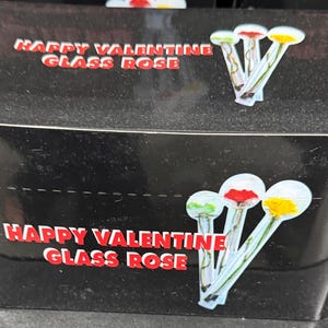 May include: Black box with the text "HAPPY VALENTINE GLASS ROSE" in red and white. The box has illustrations of glass roses with colorful tips. The roses have green, red, and yellow tips.