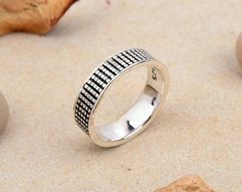 May include: A silver ring featuring a repeating black grid design around the band. The ring sits on a light brown surface, with dried leaves in the background. The ring is a simple band style.