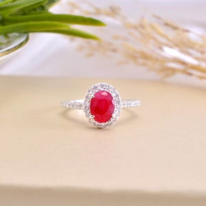 Puede incluir: Un anillo de plata con una gran gema roja brillante de forma ovalada en el centro, rodeada por un halo de piedras más pequeñas y claras. La banda también está engastada con pequeñas piedras claras. El anillo se muestra sobre una superficie de tono neutro.