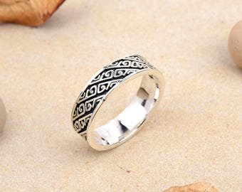 May include: A silver ring with a detailed, repeating black pattern. The ring is circular and rests on a light tan surface, with organic shapes in the background. The ring's design is symmetrical and intricate.