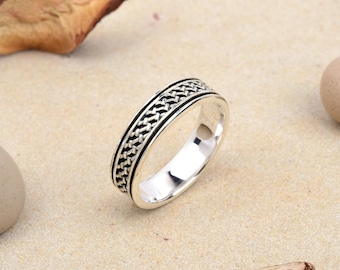 May include: A silver ring with a woven black pattern around the band. The ring is set against a neutral background with dried leaves and small stones. The ring's design is detailed and the metal is polished.