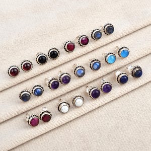 Op de afbeelding: Een verzameling oorstekers met verschillende edelsteen cabochons. De oorbellen hebben een zilverkleurige, geschulpte bezel setting. Edelstenen zijn onder meer rood, zwart, blauw, paars en wit. De oorbellen worden gepresenteerd op een neutraal gekleurde stof.