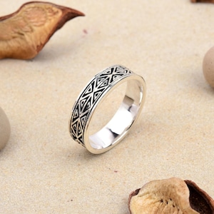 Könnte beinhalten: Ein silberner Ring mit einem geometrischen Muster aus schwarzen Dreiecken und Linien. Der Ring liegt auf einer hellen Oberfläche mit getrockneten Blättern und einem Stein im Hintergrund. Das Design des Rings ist symmetrisch und detailliert.