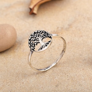 May include: A silver ring featuring a detailed tree of life design. The ring has a circular emblem with the tree's branches and leaves. The band is smooth and polished. The ring is displayed on a sandy surface.